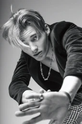 Monochrome portrait of a young man wearing a dark blazer and chain necklace, reaching toward the camera against a plain background.