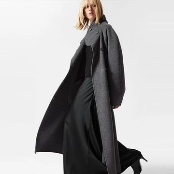 A blonde woman walks in a fashion shot. She wears a long grey wool coat, black top, and flowing pants against a white backdrop.