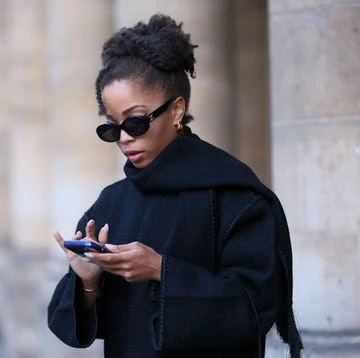 An adult female wearing a black coat and sunglasses looks at a smartphone against an out-of-focus stone facade background.