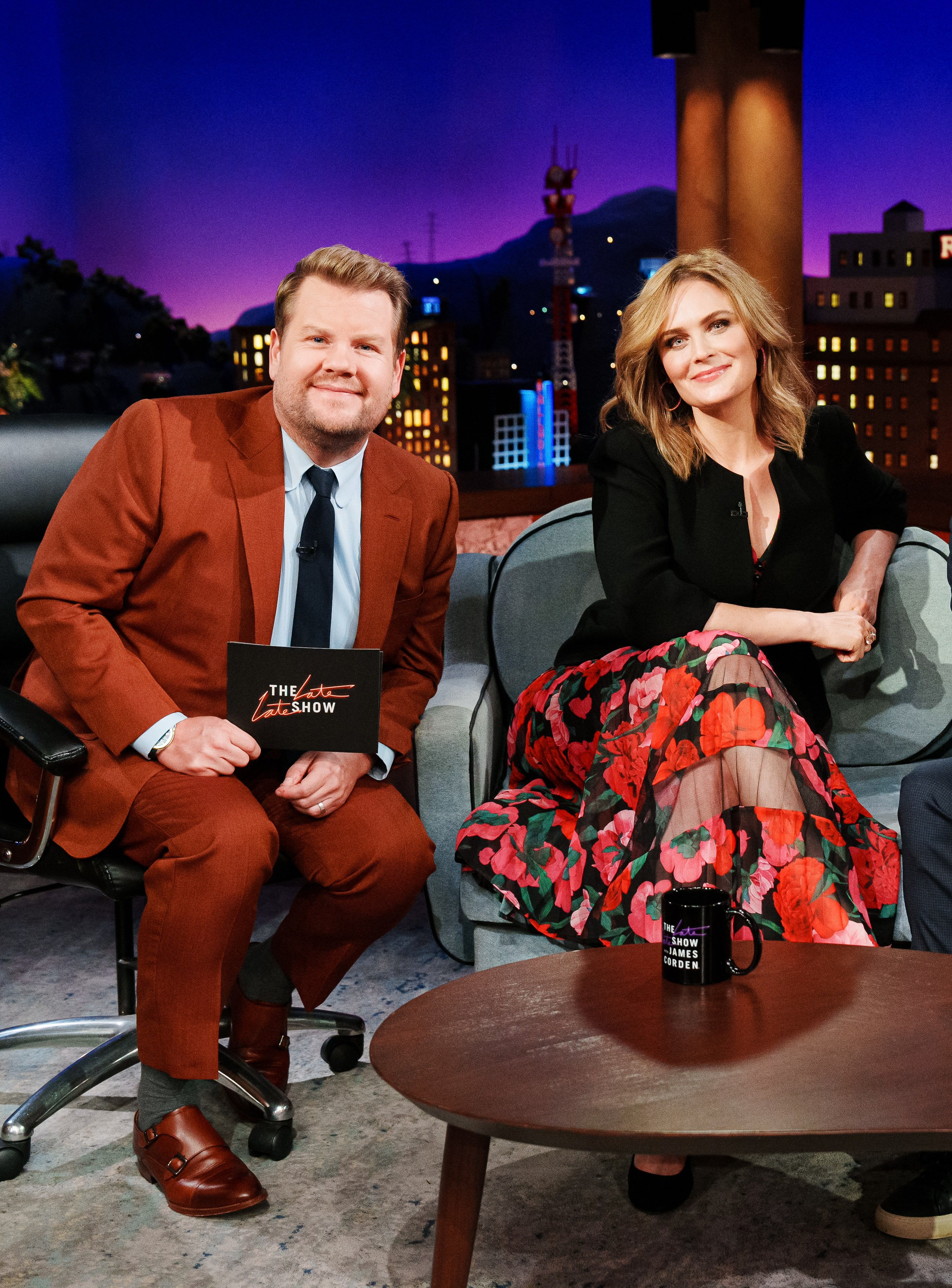 James Corden and Michelle Pfeiffer sitting on a couch in 'The Late Late Show' set with a cityscape backdrop at night.