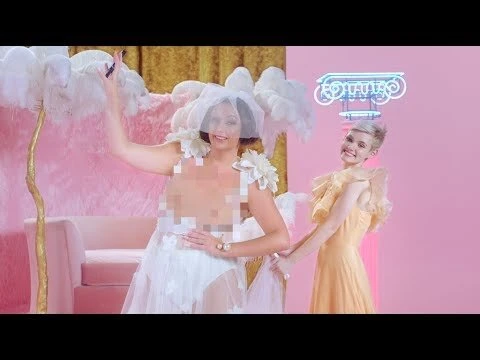 Two adult women pose in a pink studio. One wears a white dress with floral accents and a flower headpiece. The other wears a yellow dress.