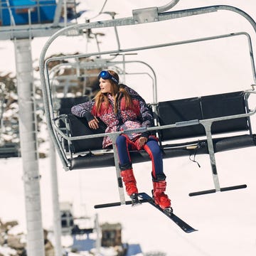 Woman on a chairlift at a ski resort with mountains in the background. She is wearing ski goggles and winter clothing.