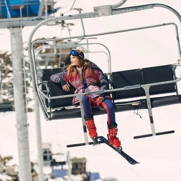 Woman on a chairlift at a ski resort with mountains in the background. She is wearing ski goggles and winter clothing.