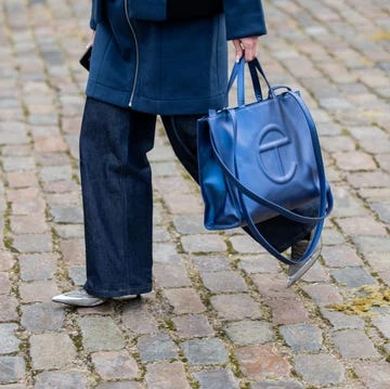Street style fashion featuring a blue Telfar shopping bag, dark denim wide-leg pants, a blue jacket, and gray pointed-toe shoes on a cobblestone pavement.