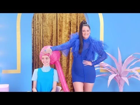 Two women pose indoors against a blue backdrop with gold curtains. One wears a blue dress, while the other has a pink headdress.