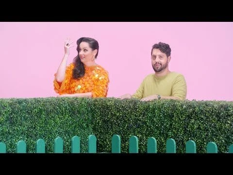 Woman in orange top and man in yellow shirt stand behind a green hedge against a pink background in this studio shot.