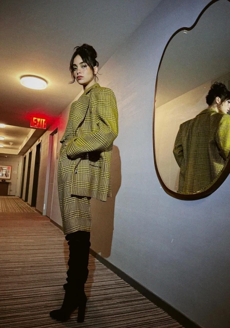 A young woman poses in a hallway wearing a plaid suit and black boots, a mirror reflecting her image; warm indoor lighting.