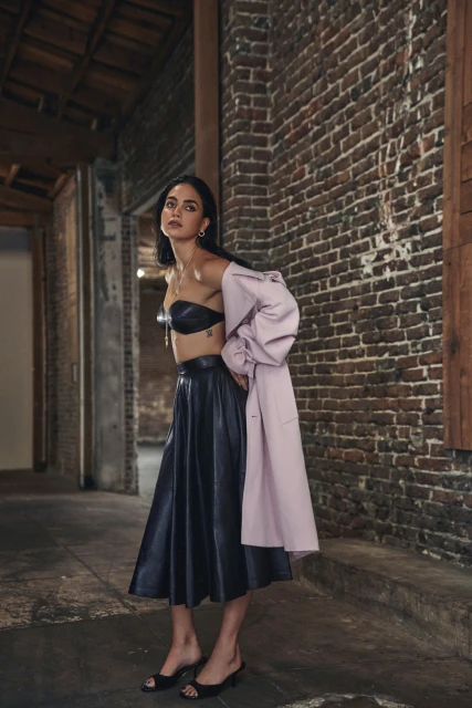 Indoor shot of a woman in a bra top and skirt with a coat draped over her arm posing near a brick wall with natural light.
