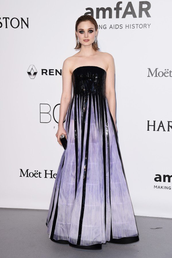 A stylish adult female attends amfAR event, wearing a black and purple gown, carrying a clutch purse, posing against a white backdrop.