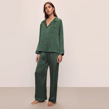 Adult female posing in full-length shot wearing emerald green silk pajamas indoors against a neutral wall in a fashion studio setting.