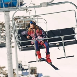 A female model seated on an open ski lift wearing patterned winter outerwear, goggles, and bright red ski boots.