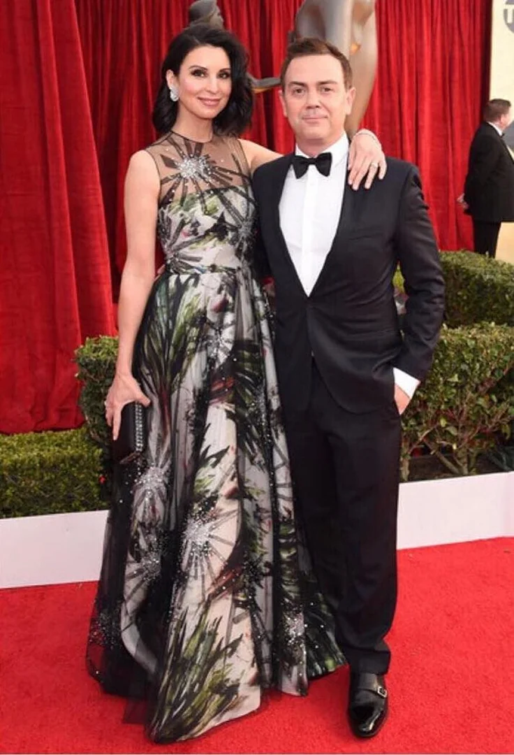 Elegant couple poses on a red carpet at an event. The woman wears a long floral gown; the man wears a black tuxedo.
