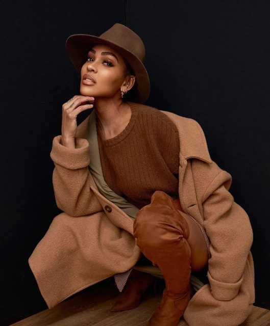 Portrait of a woman in a brown hat and coat sitting against a dark background. She has one leg raised and is looking at the camera.