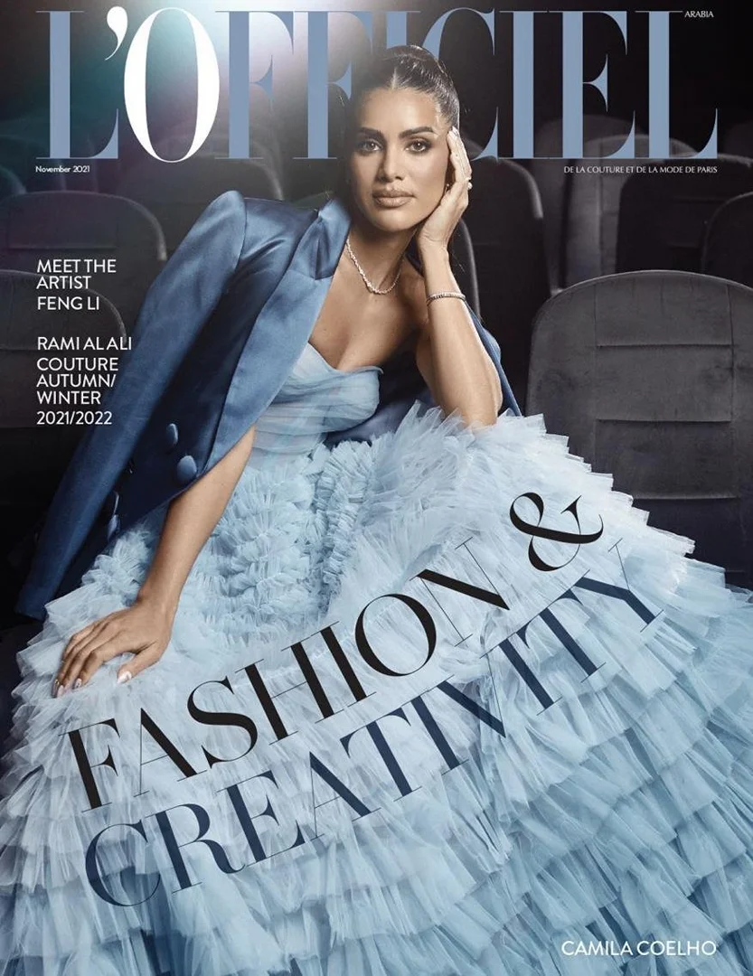 Camila Coelho graces L'Officiel Arabia cover, wearing a powder blue strapless dress and jacket, seated against theater seating backdrop.