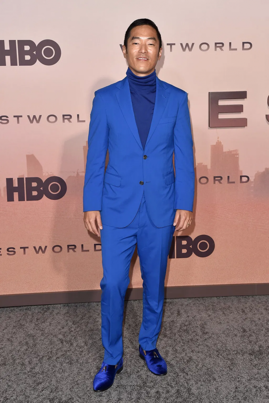 Portrait of an Asian adult male in a bright blue suit and blue turtleneck, posing on a carpeted floor in front of the Westworld backdrop.