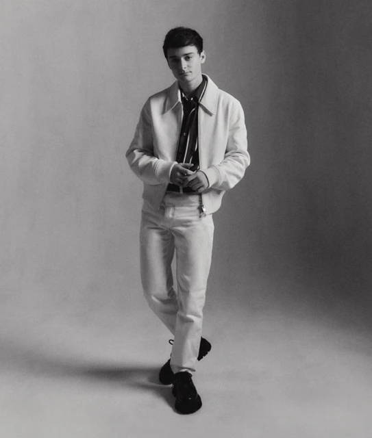 Full-length portrait of a young adult man in a white jacket and pants against a neutral backdrop, captured in a studio setting.