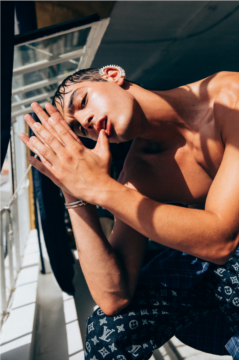 Close-up of a young adult male posing shirtless with hands together and blue pattern pants near a window with natural light.