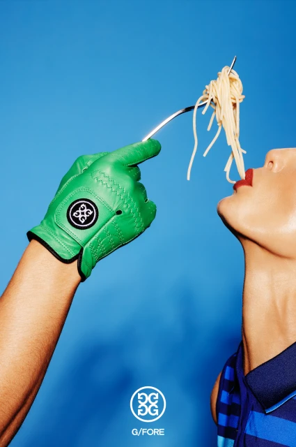 Person eating spaghetti off a fork held by a hand wearing a bright green golf glove against a vibrant blue background.