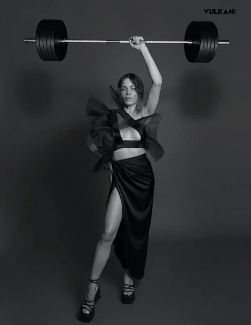 A woman in a black dress stands in front of a gray backdrop lifting a barbell overhead in a studio shot with dramatic lighting.