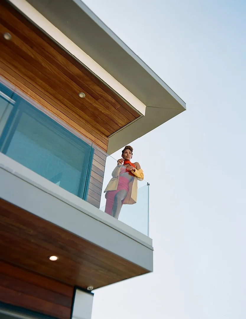 Adult woman on a modern building's balcony. Wearing a tan coat and an orange scarf, she is standing outside with a clear sky.