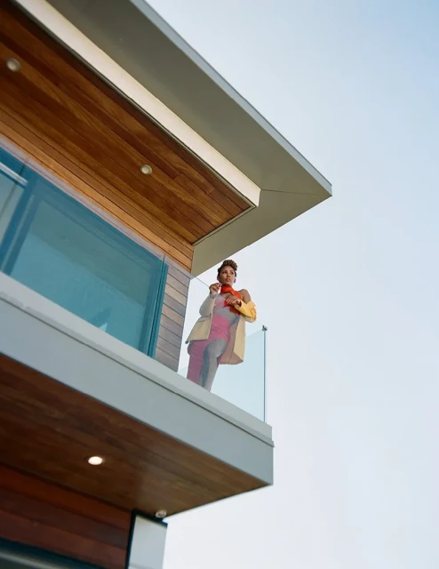 Adult woman on a modern building's balcony. Wearing a tan coat and an orange scarf, she is standing outside with a clear sky.