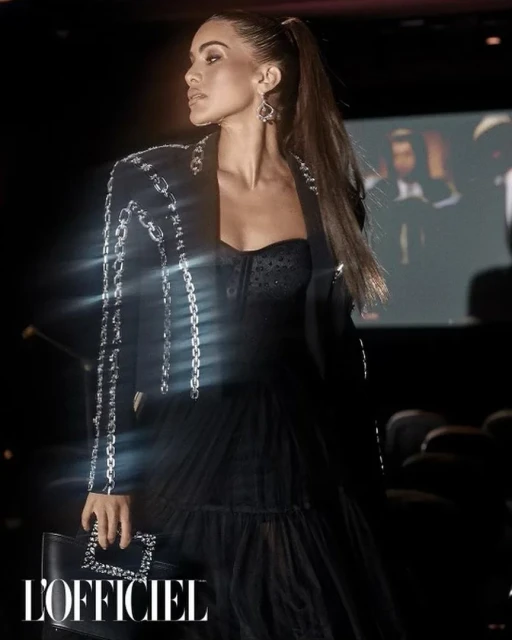 A woman poses in a black dress and jacket with silver embellishments, holding a black clutch, in an indoor setting with a dark background.