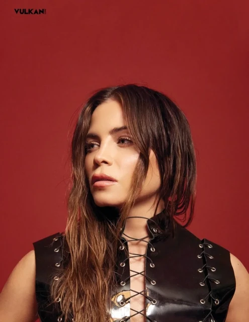 Headshot of a woman with long brown hair wearing a black sleeveless top against a red background, looking off to the left.