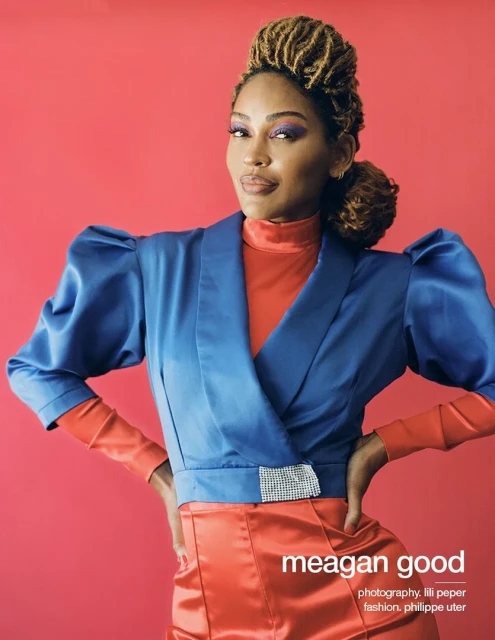 A woman with locs poses in a blue wrap jacket with orange undershirt and pants against a red background in a studio shot. She has makeup.