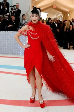 A red-dressed woman with a feathered cape poses at an event, captured in a full shot amidst a crowd and vibrant decor.