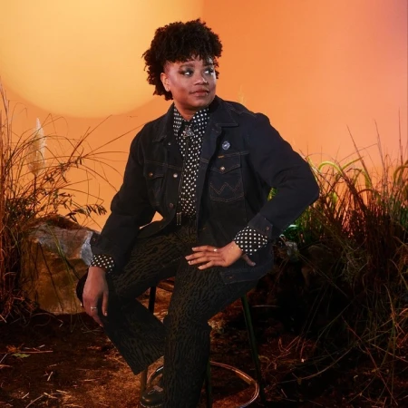 Portrait of a Black woman with short dark hair seated on a stool wearing a denim jacket and patterned trousers against a sunset backdrop
