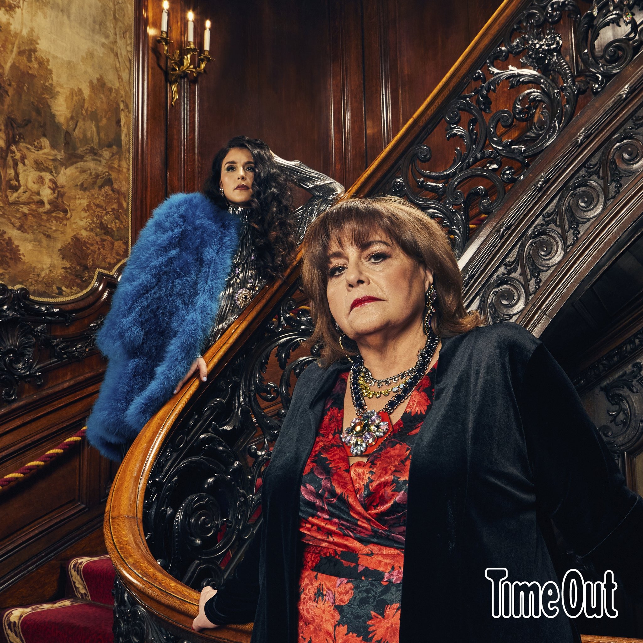 Two women pose on ornate staircase. Woman in patterned dress is foreground; woman in feathered coat is in background. Ornate wallpaper visible.