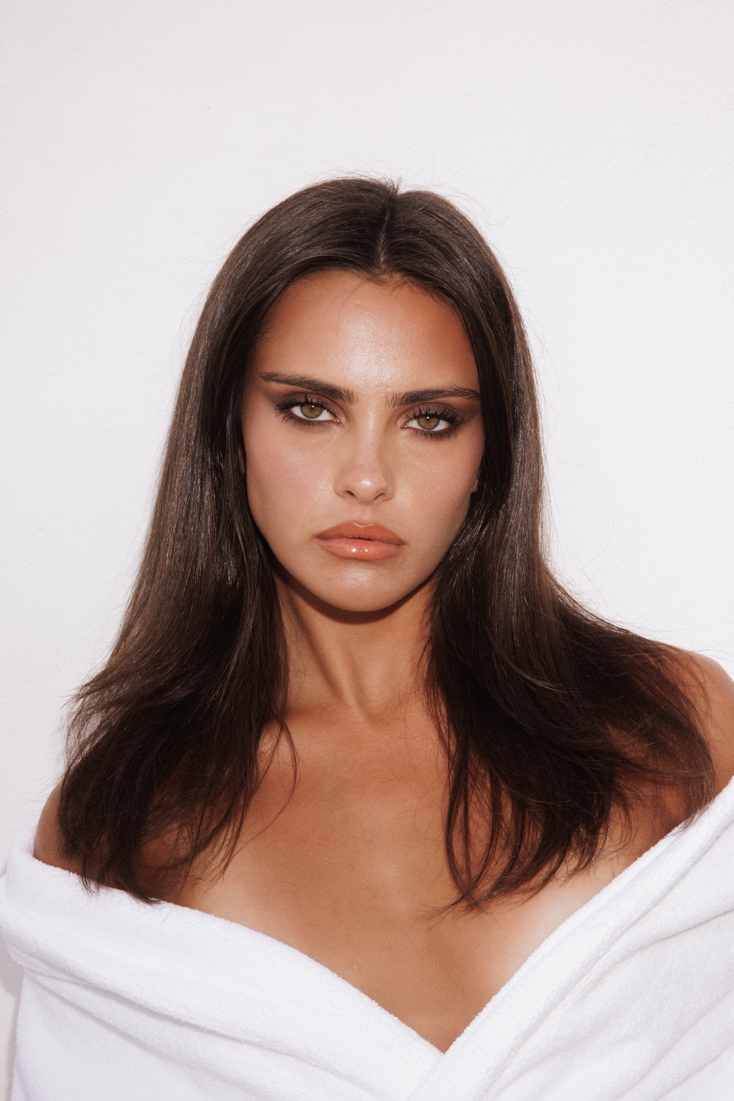 Portrait of a brunette woman with smokey eye makeup, wearing a white robe, and posing against a clean white background.