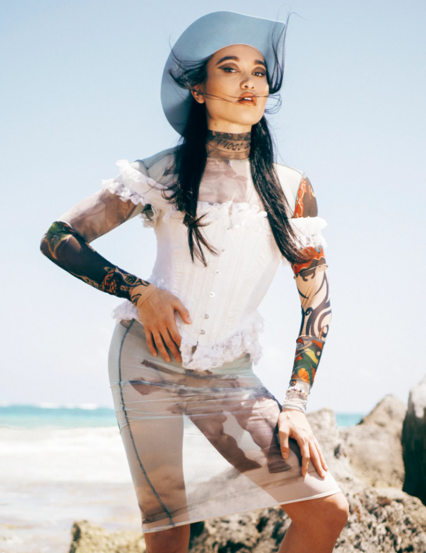 A fashionable woman poses on a beach in a white corset top, sheer skirt, and blue cowboy hat, standing in the sunshine.