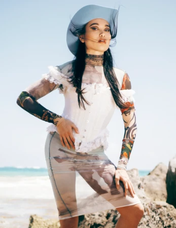 A fashionable woman poses on a beach in a white corset top, sheer skirt, and blue cowboy hat, standing in the sunshine.