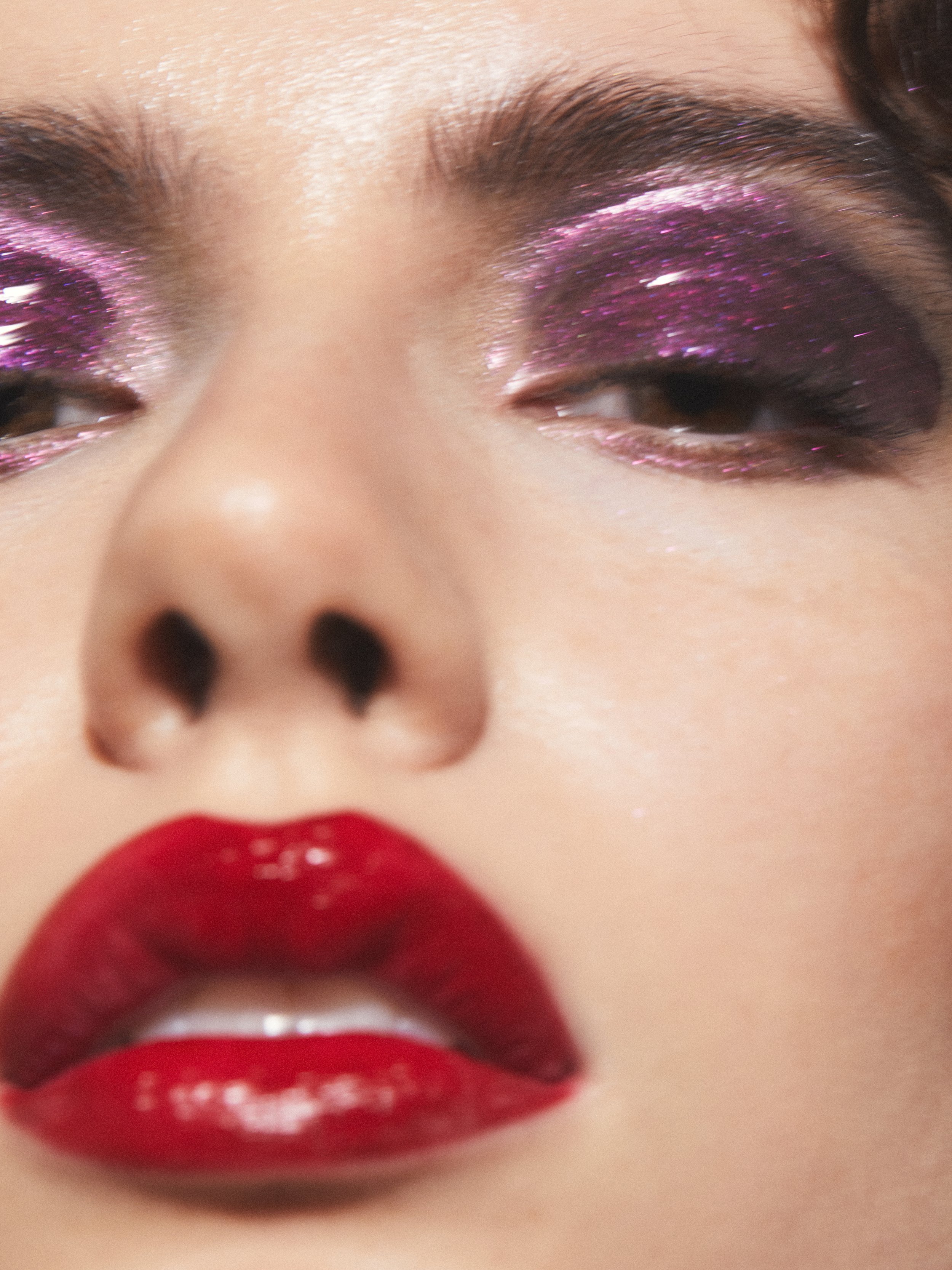 Close-up portrait of a woman's face with glossy red lips and purple glitter eye makeup in bright lighting for beauty content.