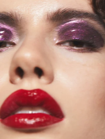 Close-up portrait of a woman's face with glossy red lips and purple glitter eye makeup in bright lighting for beauty content.