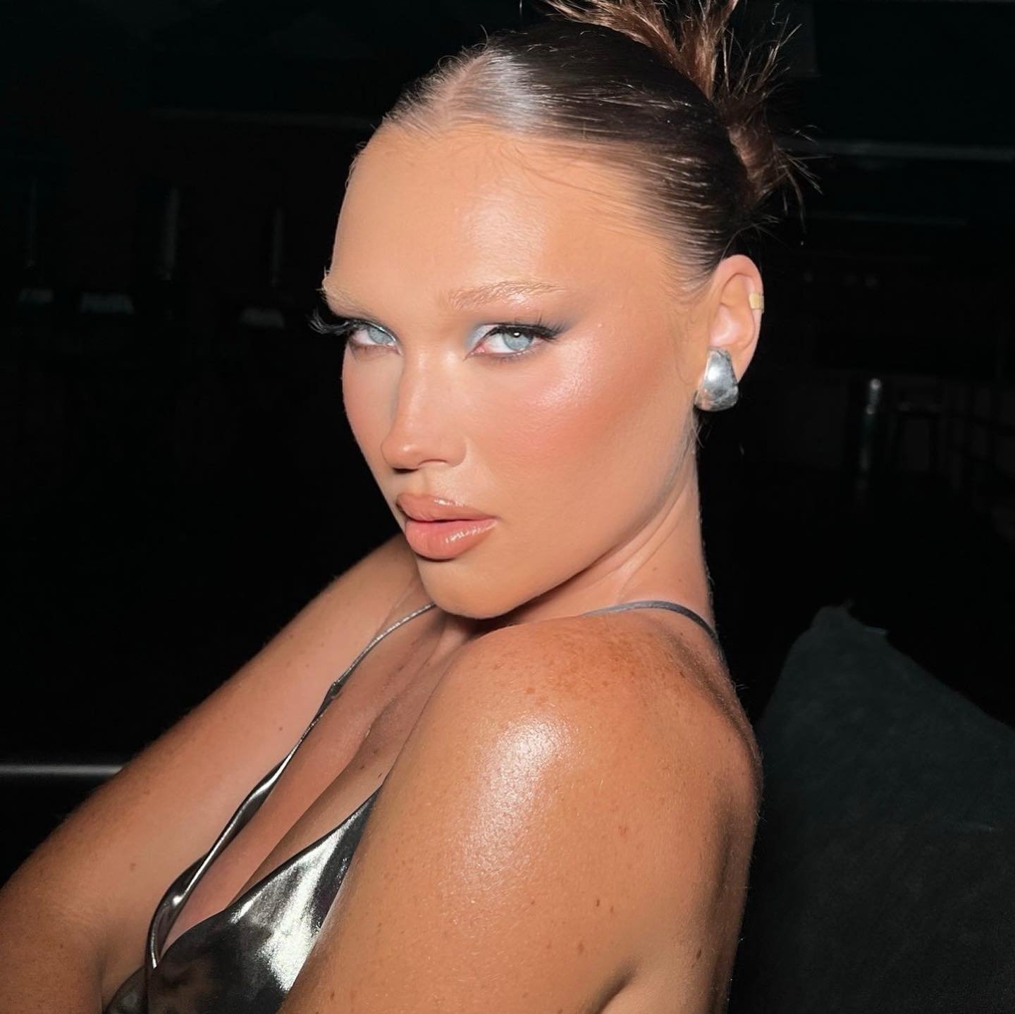 Close-up portrait of a stylish young adult woman with sleek hair, wearing statement earrings and a metallic dress against a dark background.