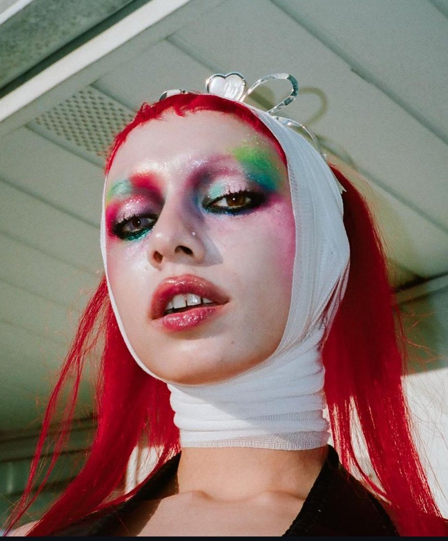 Close-up of a woman with red hair and rainbow makeup, wearing a heart-shaped crown and white bandage, gazing directly at the camera.