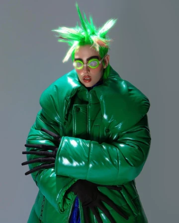 Person with spiky green hair, round green sunglasses, and a shiny green puffer coat against a grey backdrop in a studio setting.