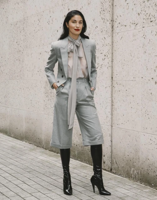 Fashionable adult woman in a grey blazer, culottes, and black boots stands against a textured wall in an urban environment.