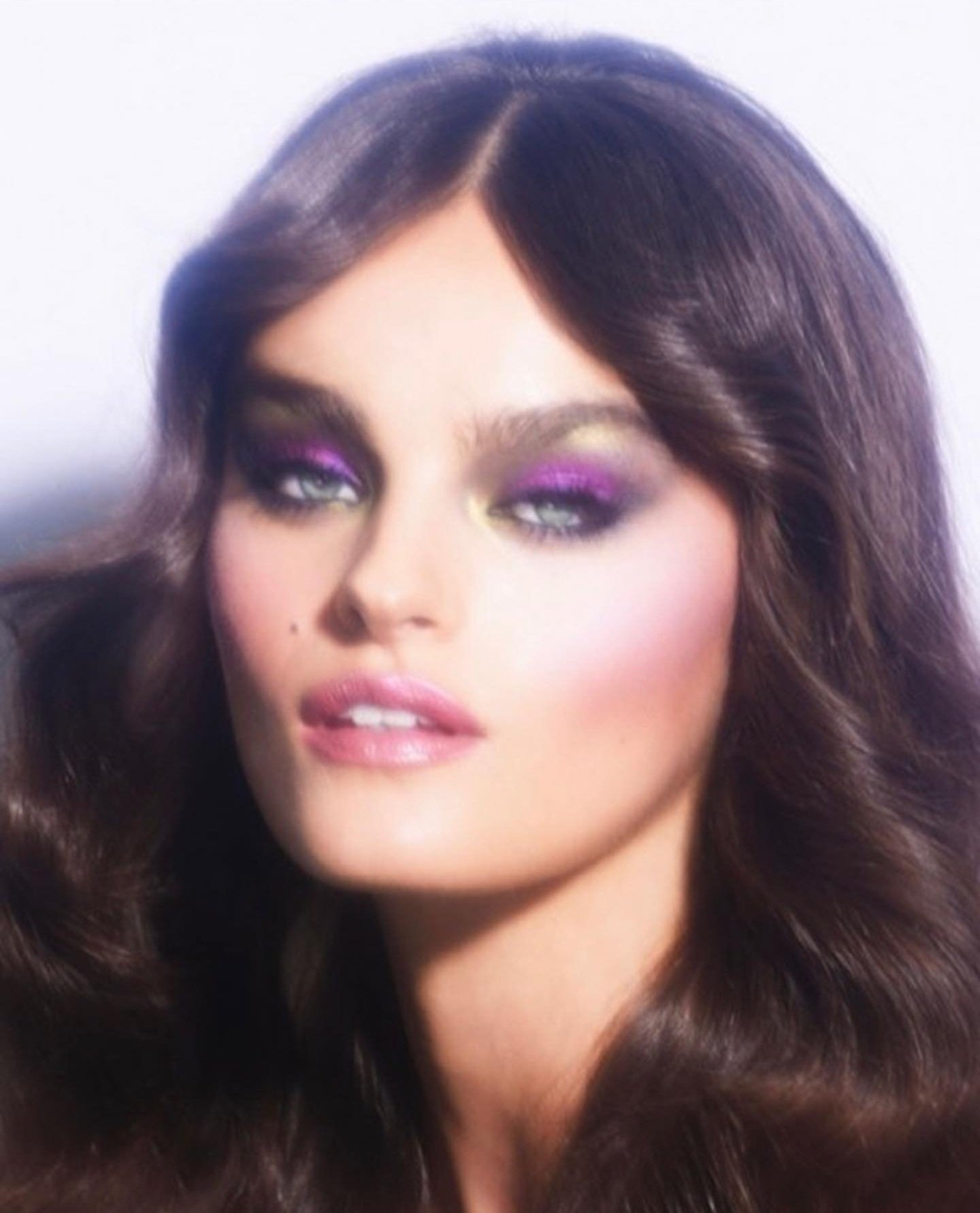 Close-up portrait of a woman with dark wavy hair and vibrant purple and gold eyeshadow, showcasing beauty and makeup artistry.