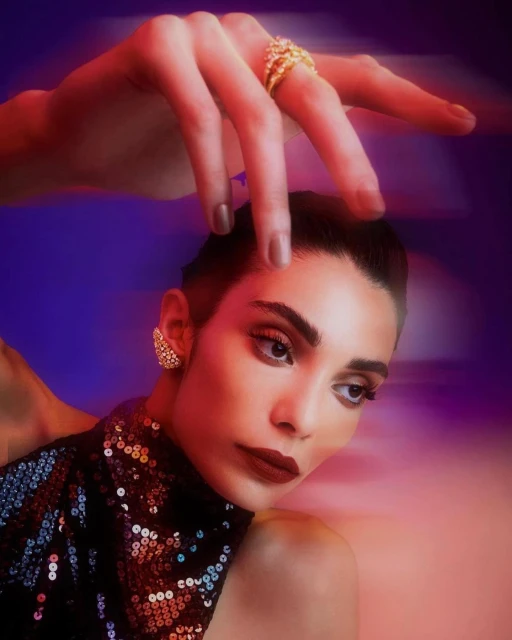 A woman with dark hair and a sequined top poses with a hand above her head, wearing a gold ring, against a blurry gradient background.