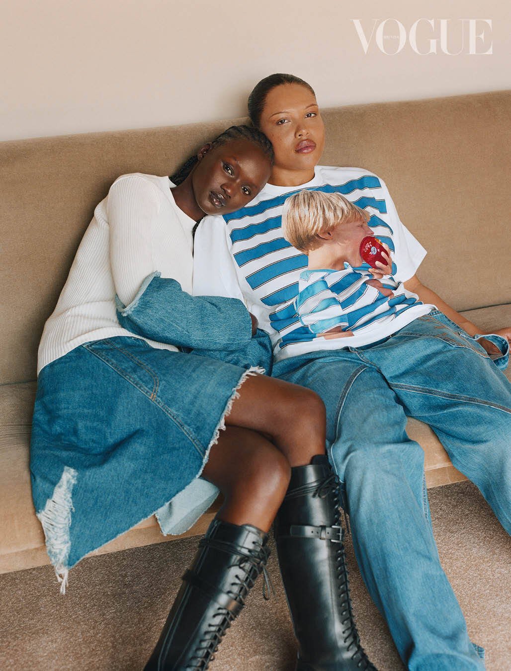 Two fashion models pose in denim and boots on a couch in a studio, in a Vogue fashion editorial with indoor lighting.