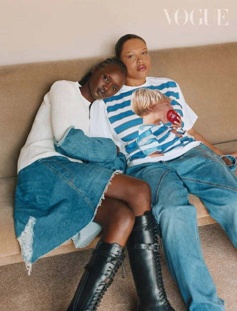 Two fashion models pose in denim and boots on a couch in a studio, in a Vogue fashion editorial with indoor lighting.
