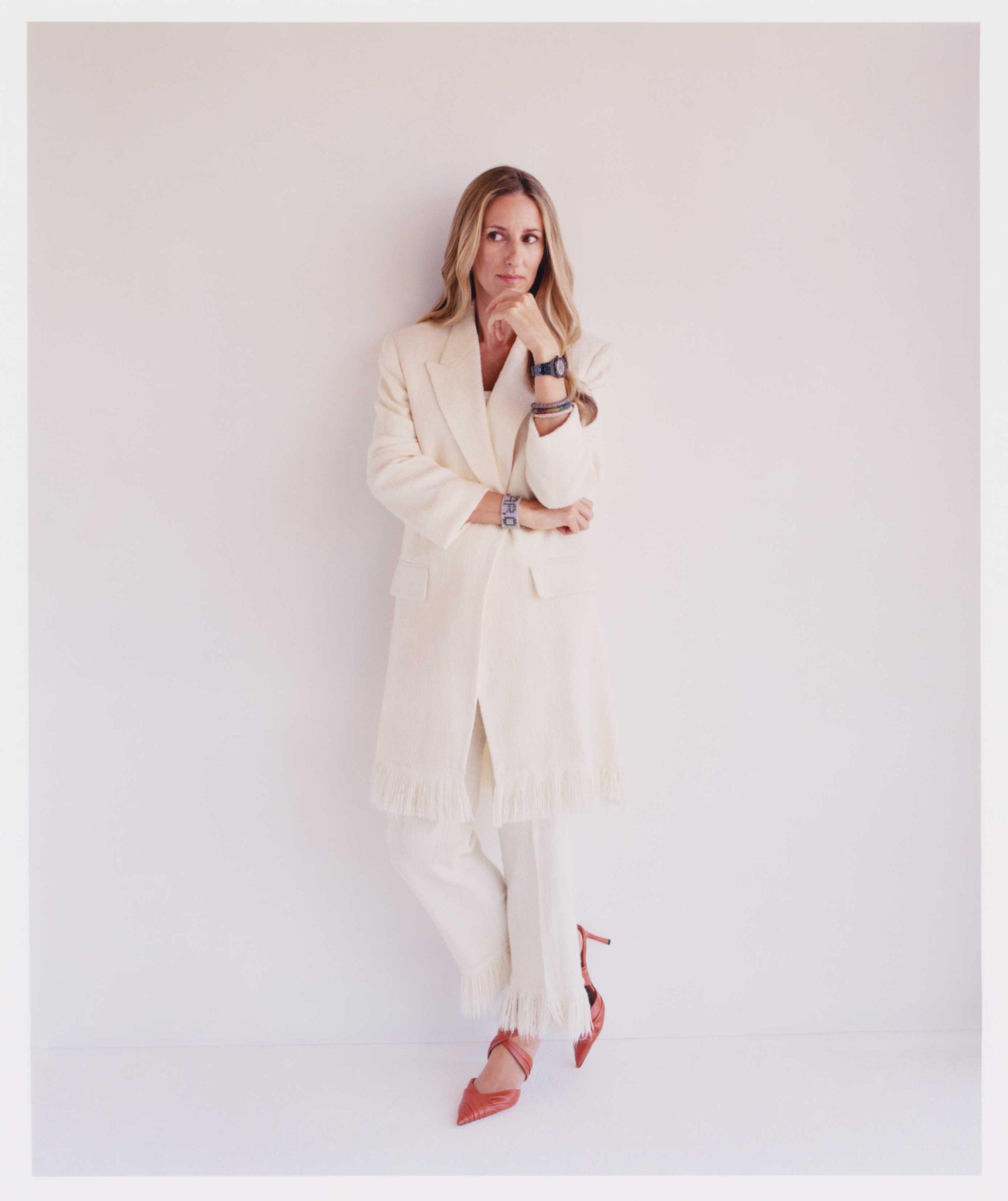 Blonde woman in an off-white suit, hand to her chin in thought, accessorized with bracelets, a watch, and red high heels in a studio setting.