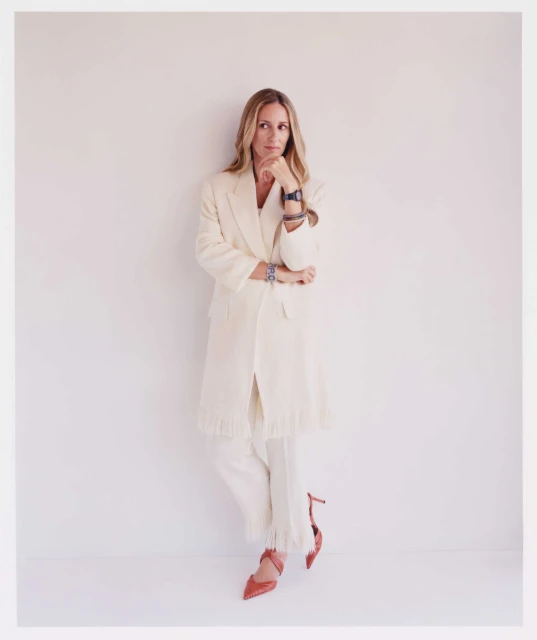Blonde woman in an off-white suit, hand to her chin in thought, accessorized with bracelets, a watch, and red high heels in a studio setting.