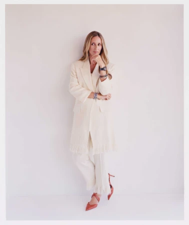 Blonde woman in an off-white suit, hand to her chin in thought, accessorized with bracelets, a watch, and red high heels in a studio setting.
