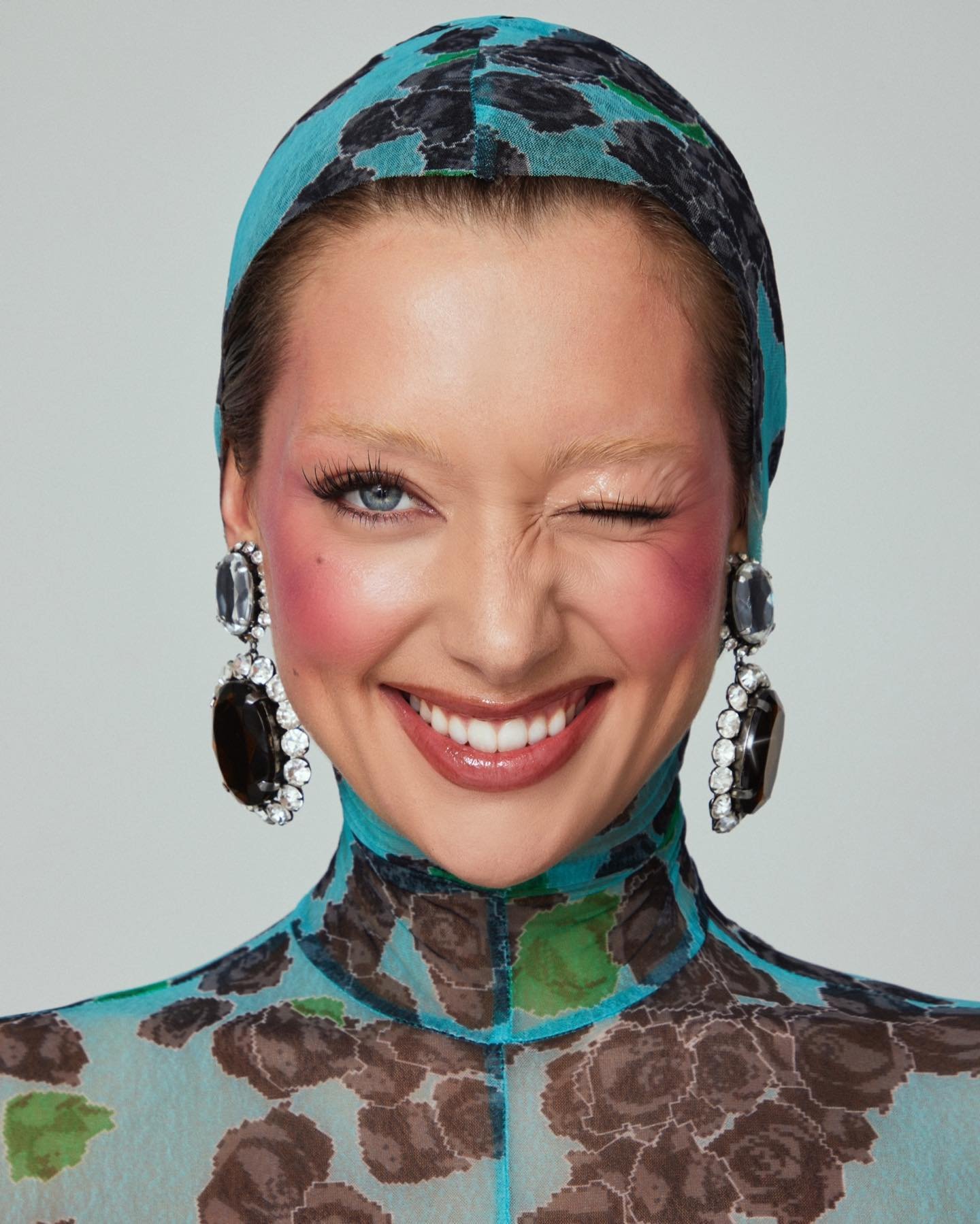 Close-up portrait of a smiling woman with pink blush, wearing a floral top and headscarf, posing in front of a neutral background.