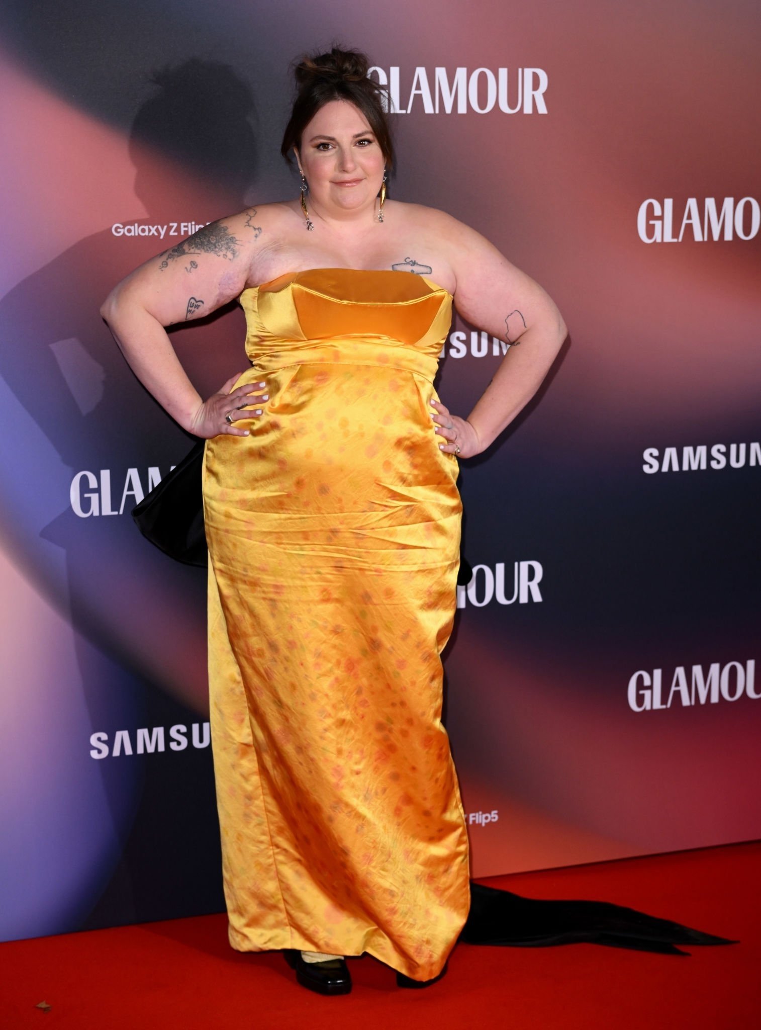 Lena Dunham on a red carpet poses in a golden strapless gown against a backdrop featuring logos for Glamour and Samsung products.