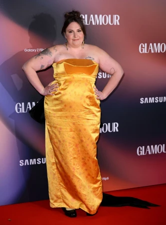 Lena Dunham on a red carpet poses in a golden strapless gown against a backdrop featuring logos for Glamour and Samsung products.
